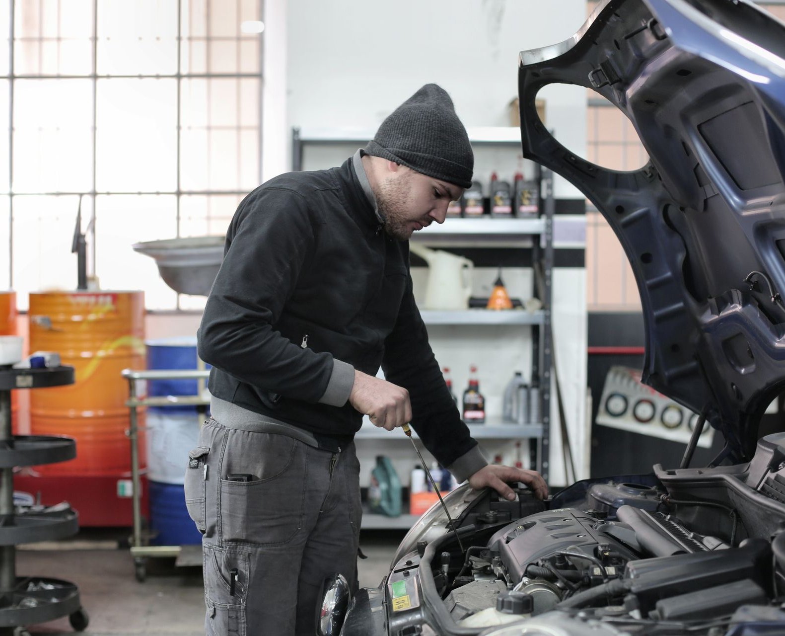 Mechanic leaning under a popped hood checking the engine bay with an oil dipstick in a repair shop